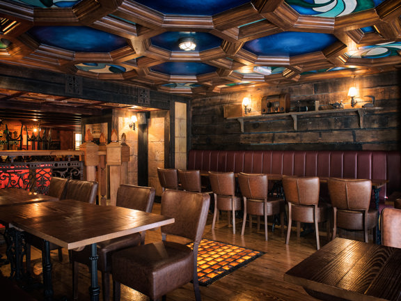 An image of Waxy O'Connor's "Cottage," semi-private dining space. The room is decorated in dark woods and with leather furnishings. The walls are panelled in rustic planks while the ceiling is ornately carved with blue stained glass details in the reliefs. Dark tables are arranged around the edge of the building against red leather benches, brown leather chairs face them. Carved posts and railings at the left end of the space separate it from further dining rooms.