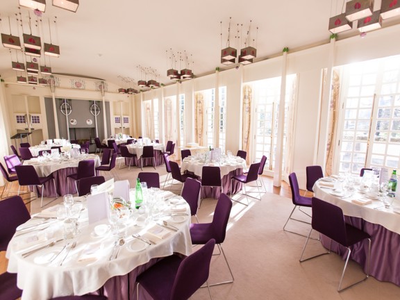 An internal view of the House for an Art Lover's Music Room laid out for a dinner. A bright, white room with beige carpet is filled with round tables covered in white table cloths. The right hand wall has floor length windows at intervals with white frames and small square panes. The chairs are deep purple velvet with silver wire bases. A small lectern is visible in the back- right corner. The light fixtures and a fireplace at the end of the room are decorated with geometric "Glasgow Style" motifs.
