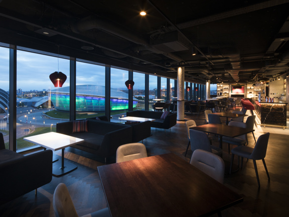 An interior view from the Radisson Red Sky Bar taken at dusk. The image shows a cosy bar with a mix of low, plush seating and tables and high velvet stools at the bar. The entire left hand wall is windowed floor to ceiling out of which the SEC campus can be seen lit up. The bulbous OVO hydro building is lit in a green-blue gradient. The bar is long and filled with modern furnishings; pendant lights add a cosy glow and golden light emits from around the bar area on the right and from the far, shelved wall.