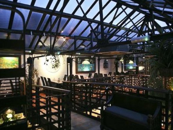An interior image of the Ubiquitous Chip's mezzanine level. A balcony space and walkway lined with black railings and a peaked glass ceiling is decorated with fairy lights, plants and paintings. Tables and chairs can be seen against the far wall. 