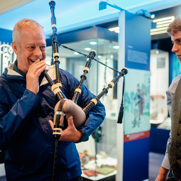 A smiling man is about to blow into a bagpipe, while a smartly dressed younger man looks on