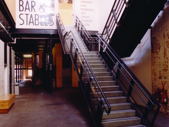 An interior view from Tramway's entrance corridor shows a post-industrial space with white-washed brick walls, a vaulted skylight roof and wooden beams. The floor has a polished concrete finish. Staircases leading off of the corridor on the right have steel tread plate and black, metal bannisters. A large wall painting on the first storey wall reads, in black writing, "Cafe bar & stables" and smaller writing lists, Exhibitions, workshops, events etc. Toilets and galleries are signposted on a pillar beyond.