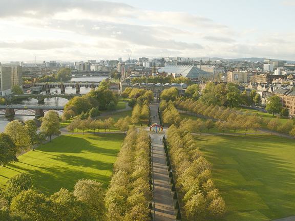 Sunny view of the tree-lined paths of Glasgow Green, on the bank of the River Clyde with 4 bridges and Glasgow city centre in the background