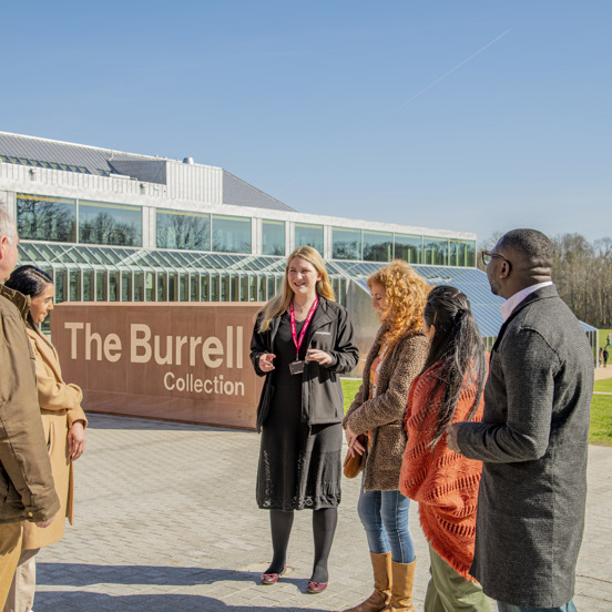 Group of people standing outside a modern red brick and glass building, a museum known at The Burrell Collection. Sun is shining, blue sky and a brick sign behind the group reads The Burrell Collection 