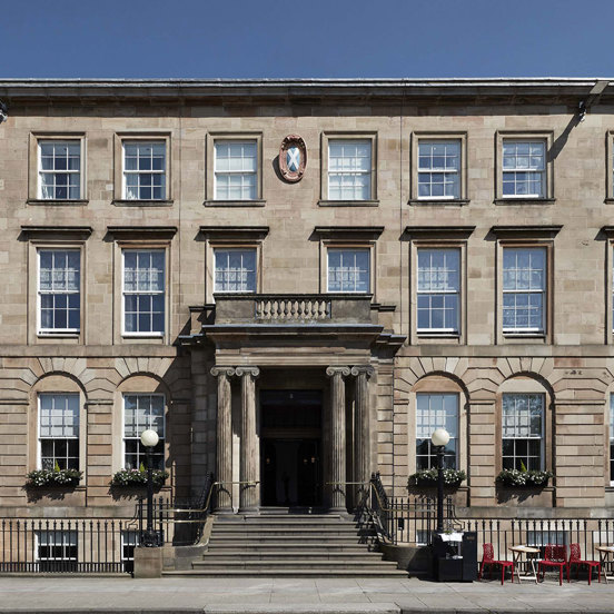 An exterior view of the Kimpton Blythswood hotel shows a grand 4-storeyed Victorian sandstone building or terrace. It is flat fronted with black railings running along its front and a grand doorway at its centre. A few steps lead up from the pavement to the doorway which is framed by Greek Ionic carved columns. The arched windows on either side are decorated with window boxes, 2 black iron, ball-topped lampposts adorn the entrance further. Small bistro tables and chairs line the pavement on the right.