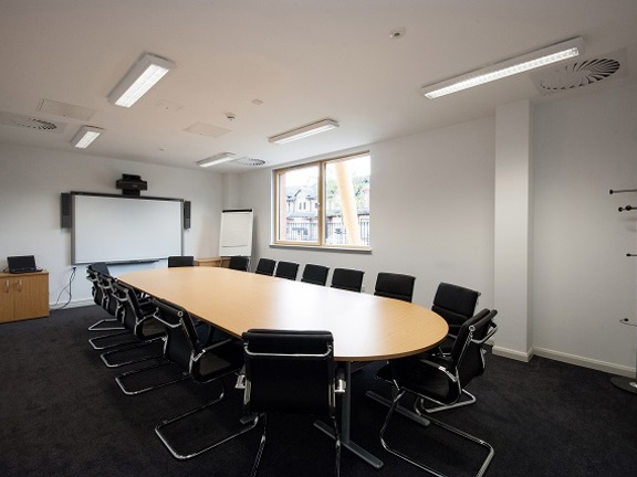 A photo of a board room style meeting space at the William Quarrier conference centre. The room has a dark grey carpet and white walls. A large, oval, pale wood-effect table sits in the centre of the room surrounded by black padded chairs with chrome bases. The end wall has a smart board mounted on the wall, a small monitor sits on a low sideboard to the right. An A-frame, window and coat hook can also be seen in the room. Florescent lights and vents can be seen in the ceiling. 