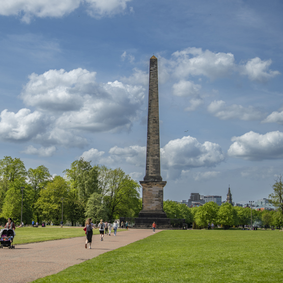 View of the 44m tall Nelson Monument obelisk in Glasgow Green, with people jogging, strolling and lounging in the sunshine