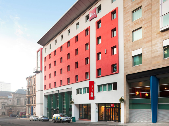 An exterior, daytime view of the Ibis Hotel shows a 7-storey modern building with square windows, on a sloping street. The building is rendered in white with red cladded details. An angled metallic roof is also visible. The entrance is a glass double door with dark frames at the right of the building. Red signs reading "Ibis" and "Ibis Hotel" respectively are visible on the building. Modern and Victorian stone building can be seen neighbouring the hotel and on the perpendicular street on the left.