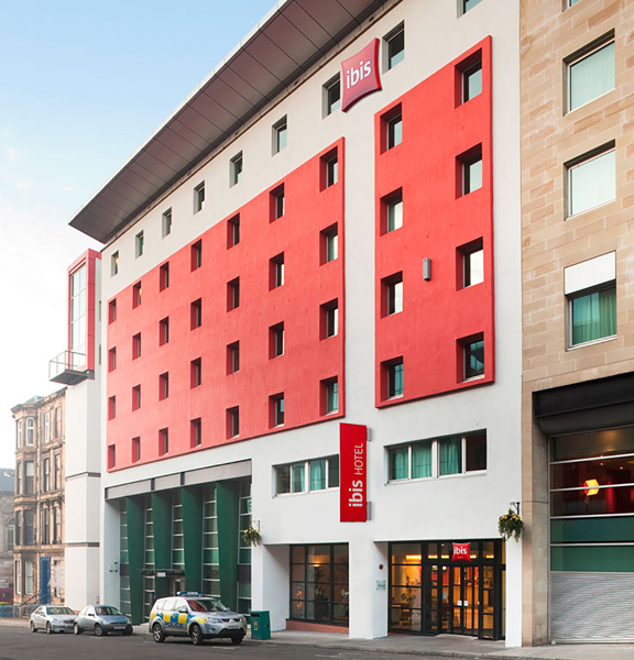 An exterior, daytime view of the Ibis Hotel shows a 7-storey modern building with square windows, on a sloping street. The building is rendered in white with red cladded details. An angled metallic roof is also visible. The entrance is a glass double door with dark frames at the right of the building. Red signs reading "Ibis" and "Ibis Hotel" respectively are visible on the building. Modern and Victorian stone building can be seen neighbouring the hotel and on the perpendicular street on the left.
