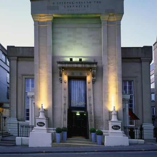An exterior image of the Malmaison Glasgow shows a sandstone building with a strikingly, tall Egyptian-influenced façade. An enormous, carved archway houses the entrance, a black double doorway with a large window above it. Gold lettering above the door in the signature branding reads, "Malmaison." The façade is uplit with white and green lights, exaggerating its height and showcasing the carved pillars and a Greek inscription at the very top. Black railings and potted shrubs decorate the steps.