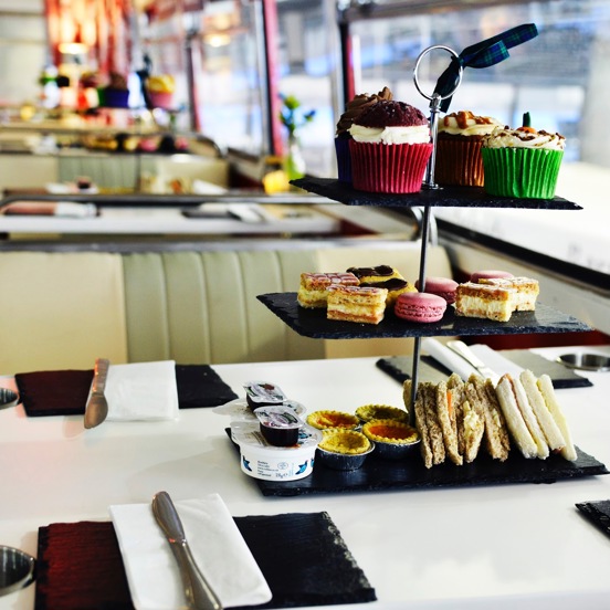 An close up image of an afternoon tea on the Red Bus bistro. A black slate cake stand with 3 tiers is adorned with small cakes in colourful paper cases, triangular, filled sandwiches and macarons. 4 places, with slate plates, knives and paper napkin are set around it. Chrome cup holders are set into the top of the while table top. The vintage leatherette bus seat with chrome edges and another decorated table can be seen to the left of the long right-hand windowed wall.