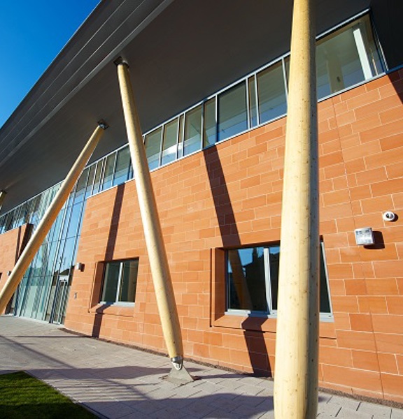 A photo looking up at the William Quarrier Conference Centre's exterior. A 2-storey modern red stone and glass building with cream, narrow, crisscrossing pillars. The walkway outside and around the building is paved with grey stone. 2 small patches of grass can just be seen either side of the paved walkway. The edge of a carpark can just be seen beyond the far-right corner of the building. 