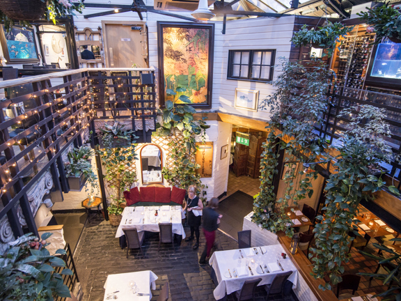 An interior image of the unique Ubiquitous Chip's main dining room, looking down from the mezzanine level above. Black railings around the balcony level are strewn with fairy lights, hanging baskets and trailing plants. The floor of the ground floor dining room is cobbled with dark stone, another section visible off to the right is tiled. Dining tables are laid with cloths, napkins, glasses and cutlery. Eclectic décor, including large paintings, fish tanks and stone carvings adorn the space.