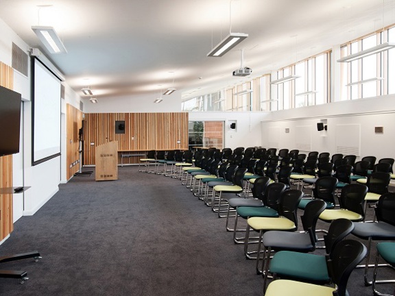 A large lecture room at the William Quarrier conference centre shows a bright room with dark carpets; the walls are painted white with wooden, slatted accents. Stacking chairs with wire legs and padded seats in shades of green and grey are arranged into 4 long rows. The chairs face the front of the room where a large projection screen, a lectern, 2 TVs on wheeled stands are visible. A projector and fluorescent lights are mounted to the ceiling & speakers dotted around the rooms walls. 