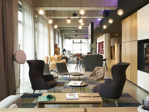 An interior view of the Moxy Merchant City lounge and reception area shows a bright, modern open plan space with glass, exterior walls. The floor is polished concrete, large rugs are placed in the seating areas. Eclectic, modern armchairs and coffee tables are dotted through the closer half of the space. A large TV can be seen mounted on a feature wall on the right. Beyond that a high counter painted grey is surrounded by high, dark metal stools; and beyond that more seating and shelves can be interpreted.