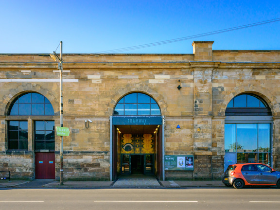 A photo of Tramway taken from over a road. A long, flat roofed Victorian, blonde sandstone building, with a huge central arched doorway and windows of the same scale on either side. The doorway has a grey sign reading "TRAMWAY" and set back from the street - above an automatic glass door - is a large yellow art work taking up the height of the entrance way. A parked car, bike racks, a lamppost and a CCTV camera can all be seen. The pavement is tarmac but the entrance to Tramway is paved with bricks.