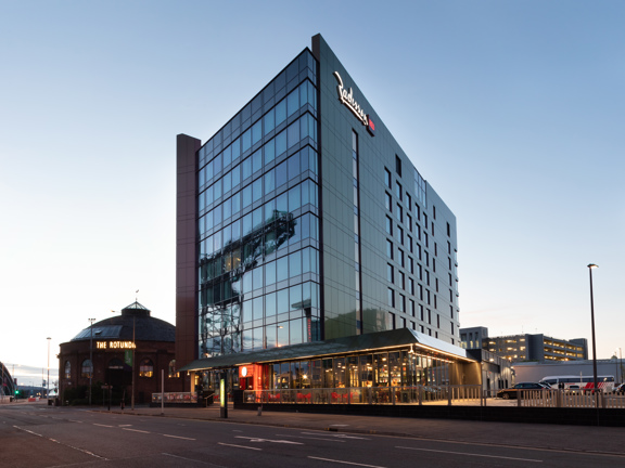 An exterior image of the Radisson Red hotel taken from over a road. A modern multi-storey building with a dark windowed front and dark grey, metallic cladding on the sides. A large lit-up sign of the Radisson Red logo adorns the top portion of this side wall. A narrow pavement runs the length of the road, off of which a few stairs lead to an entrance. A glass railing separates the pavement from a terrace with seating and a car park. At the end of the road buildings, including the SEC Armadillo are visible,