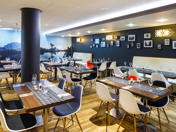 An interior image of the Ibis Hotel restaurant, the floor is a light wood, the left hand wall is covered with a photographic image of the SEC campus reflected in the River Clyde, the other is painted dark blue; a cylindrical pillar in the centre of the room is painted the same shade. Wooden tables of 4 to 8 settings dot the space. Those against the walls have plush white benches, the other chairs are white, formed plastic with grey padding and teak legs. Framed prints and lights decorate the dark wall.