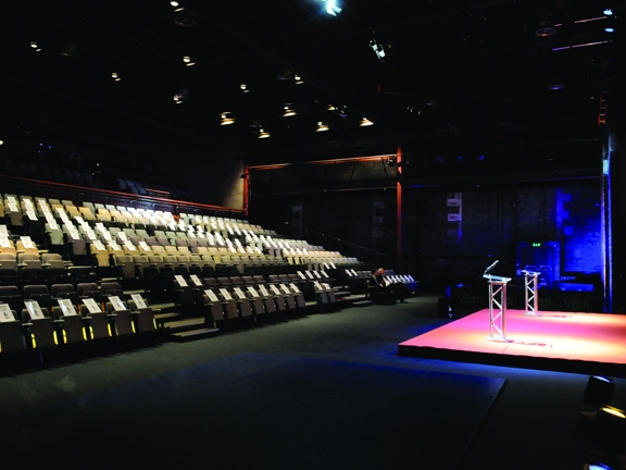 A view of the Tramway Theatre from the level access entrance. A large theatre with dark floors and walls. To the left, the majority of the room is filled with rows of tiered, self-right seating. To the right a small stage with two metal lecterns is lit with coloured spotlights. A large projection can also be interpreted o the wall above.