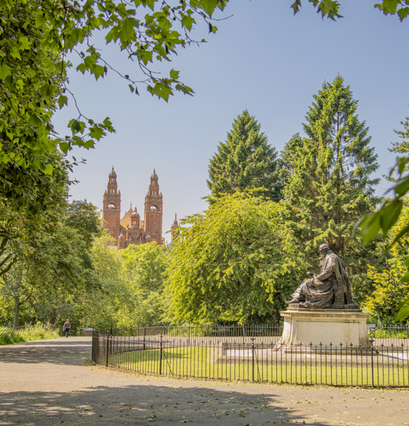 View of a large, sitting statue of Lord Kelvin, surrounded by green trees in Kelvingrove Park, with the towers of Baroque-style Kelvingrove Art Gallery and Museum in the background