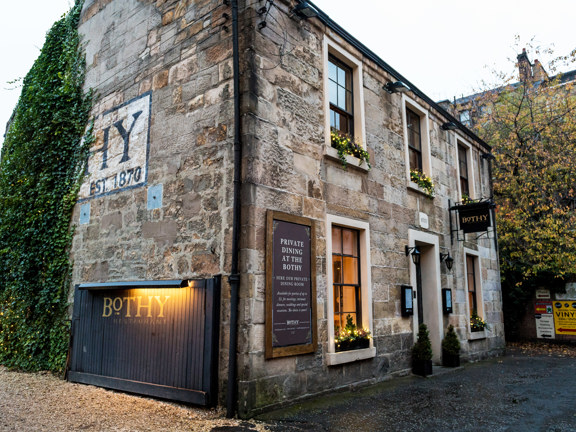 Exterior image of The Bothy restaurant shows an old, 2-storey sandstone building. The front of the building has traditional windows with dark frames, window boxes and small potted trees decorate the windows & doorway. The side of the building is half covered in ivy, obscuring a large wall painting, the "HY" of Bothy and "est.1870" are visible. A wide wooden gate, propped open against the building and a black hanging sign above the door have gold lettering reading "Bothy". The ground is paved and gravel. 