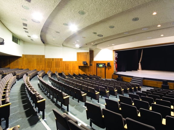 An interior view of a theatre-style space at the Mitchell Complex, a large, curving, carpeted auditorium with stepped seating. The ceiling has a textured white rendering and the walls have golden coloured wood panelling or tiling applied to the lower half of the walls and painted white on the top portion. The carpets are grey and the brown, fabric rows of chairs have chairs have retracting seats. At the far end of the room a large stage, with stairs up, technical lighting and curtained wings can be seen. 