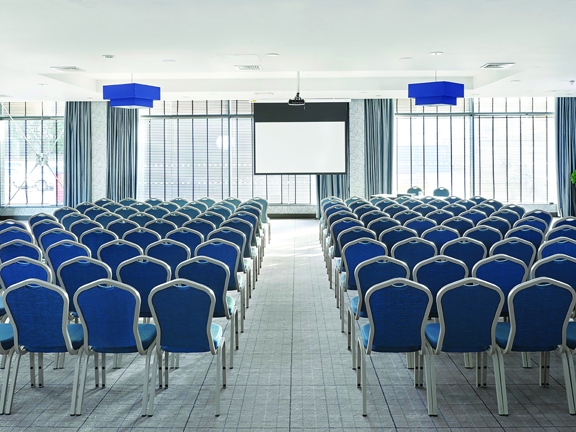 A photo of the Village Hotel's Meeting Room shows a bright room with pale. stripped carpets and 2 glass walls. The windows are covered in open, venetian blinds, pale blue curtains are also pulled back. The room is largely filled with rows of turquoise chairs with metal legs, at the front of the central isle, a projection screen is pulled down from the ceiling - where a projector is also mounted. Royal blue lampshades, small potted topiary plants and a framed print further decorate the room.