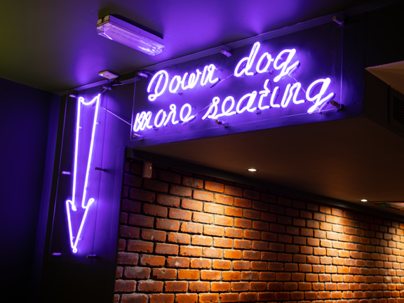 A blue neon sign hangs in a bricked corridor. It shows a large downward pointing arrow and reads "Down dog more seating" in a cursive style.