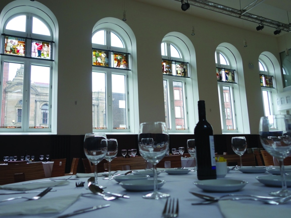 A view of the Maryhill Burgh Hall arranged for a dinner event. The photo is taken from table-level, it's possible to make out multiple tables - laid with silverware, glasses and bottles of wine. Wooden-backed chairs surround the tables. The wall is half panelled with dark wood, the upper part is painted white. 5 large arched windows are visible in the wall, stained-glass panels depict people of different trades and professions. A technical lighting rig and florescent lights can be seen on the ceiling.