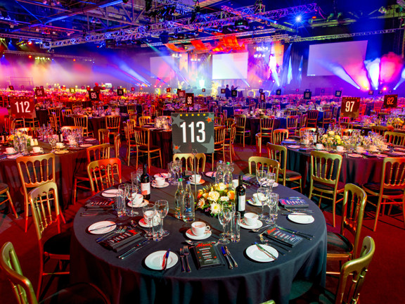 An SEC interior arranged for a large private dinner. A large exhibition hall is filled with large round tables with black tablecloths, surrounded by gold chairs. The tables are numbered (113 is closest) and decorated with cut flowers, menus and silverware. The room is primarily lit with coloured spotlights in red and blue and yellow; the technical lighting rig is visible across the ceiling. Three large, white projection screens are visible on the right wall of the space, though nothing is projected on them.