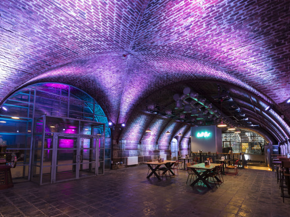 An interior image of Platform shows the junction between 2 cavernous, arched brick tunnels. The tunnel on the left has a glass and steel double doorway built into the archway., the walls on the other side are lit pink. The tunnel to the right has a tiled floor, all of the the curved walls are uplit with bright, mauve lights. Industrial-style pendant lights hang above wooden tables and wire framed chairs. A food or drink outlet at the end of the space has a turquoise neon sign on a free standing wall.