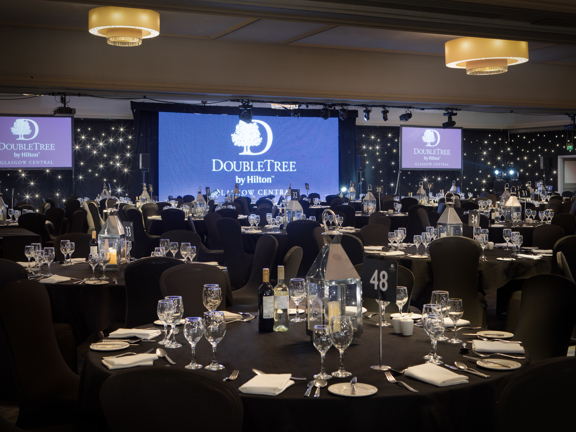 An interior view of the Doubletree by Hilton ballroom, a large room decorated with black, starlit walls. The floor is filled with large round tables, covered with black table cloths and chairs with black fabric covers. The tables are decorated with lanterns, glasses and silverware. At the end of the room there is a projection screen and a large monitor on either side, all 3 read "Doubletree by Hilton". There are chandeliers in the ceiling, a projector, a technical light rig and speakers all visible. 