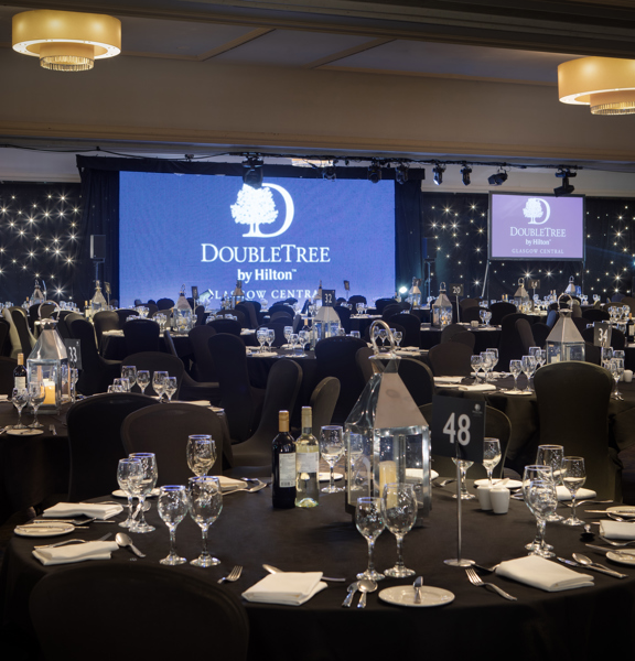 An interior view of the Doubletree by Hilton ballroom, a large room decorated with black, starlit walls. The floor is filled with large round tables, covered with black table cloths and chairs with black fabric covers. The tables are decorated with lanterns, glasses and silverware. At the end of the room there is a projection screen and a large monitor on either side, all 3 read "Doubletree by Hilton". There are chandeliers in the ceiling, a projector, a technical light rig and speakers all visible. 