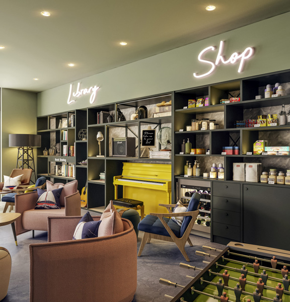 An internal view of the Adagio Hotel's lobby. The room has a large window and floor to ceiling shelves full of books, objects and supplies. The words "Shop" and "Library" are on the wall in neon. There is modern furniture and a foosball table visible.