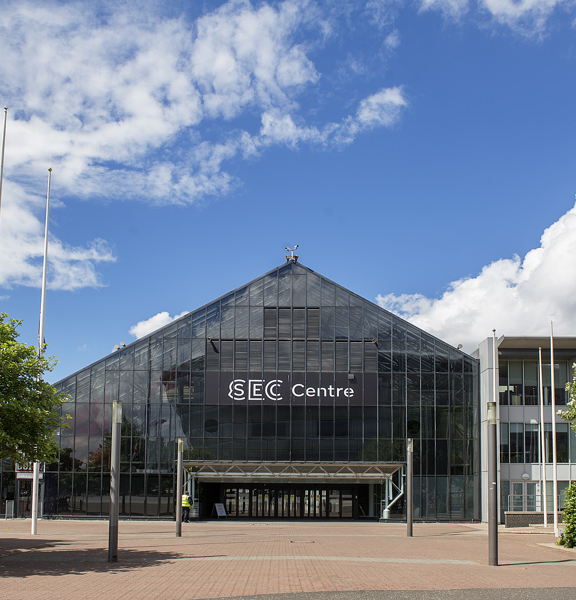 An exterior shot of the SEC Centre shows a large, glass, flat-fronted, triangular building; the facade bears a large black sign with white lettering that reads "SEC Centre". The expansive, pedestrianised space outside is paved with bricks, trees, hedges, lampposts and 3 flagpoles are visible around the buildings entrance. The building is pictured against a blue, cloudy sky.