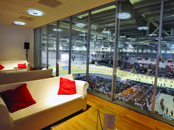 An interior image from the Emirates arena shows a room with a large plate glass wall overlooking the main velodrome arena below. The room has wooden floors and white walls. 3 white, plastic seats formed to resemble sofas are visible, with red cushions and low tables also decorating the space. A black speaker on a stand is visible in the back right corner.