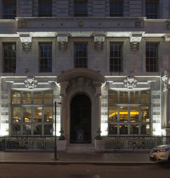 An exterior photo of The Anchor Line taken at night from across the road, depicts the first 3 floors of a grand, white Victorian building with large traditional windows.  The building is adorned with carved and gilded details & boasts 3 large carved doorways. The right hand one is lit and has dark lettering that reads "Anchorline" on its lintel. Through the window a bar can be seen lit up. The pavement outside is paved, street signs, lampposts and a parked car can be seen outside.