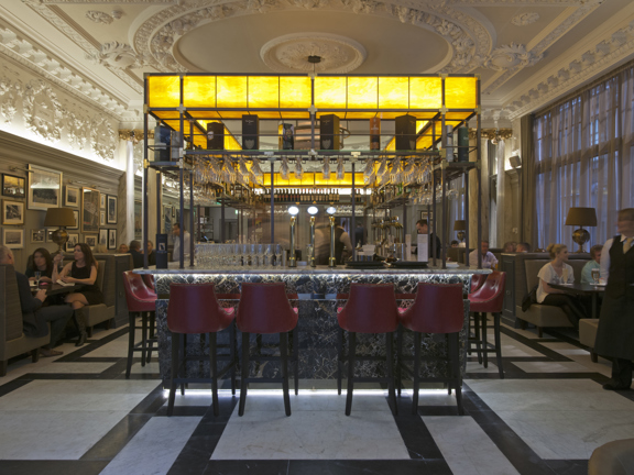 An interior view of the Anchor Line bar shows a room with a high, ornately plastered ceiling and a polished black and white marble floor. Booths with low, plush grey benches and dark tables line either wall. In the centre of the room a 4 sided, veined marble bar is lined with high, red leather stools. The bar has a dramatic, metal framework above it holding glasses and whiskey boxes. A huge golden, rectangular light fixture tops the structure on all sides. A large window can be seen in the right hand wall.