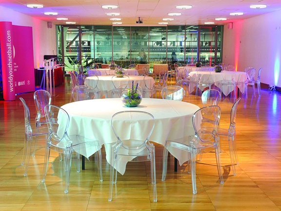 An internal view of the Emirates Arena Function Suite shows a brightly lit, white room with a polished wooden floor. There are a number of large round tables, covered in white table cloths, surrounded by transparent acrylic chairs. The far wall of the room is glass and looks out onto the arena's interior. On the left of the room a spider frame banner, a podium and a rectangular table can be seen. Speakers, projectors and plug outlets are also visible.