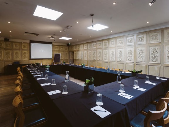 An interior view of a meeting room at Corinthian Club. 4 long rectangular tables, covered in black table cloths, are arranged into a rectangle; with wooden chairs lining the outside edge. Plants, bottles of water, paper and pens are visible on the tables. The room has a wooden floor and panelled walls. There is a large projection screen and a flipchart visible at the far end of the room. Speakers and ceiling lights are also visible.