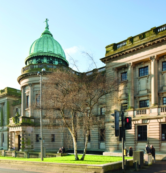 An exterior view from the street outside the Mitchell Library Complex. Shows a pedestrian crossing outside a large 3-storey sandstone institution. A verdigris dome topped with a female figure, adorns the central curving portion of the building, above a large, pillared doorway. In the foreground, a grassy bank with a silver birch in its centre sits slightly raised from the tarmac pavement around it.  Lampposts and a modern building can be seen in the background.