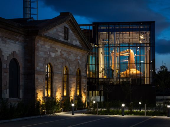 An exterior shot of the Clydeside Distillery at night, the image is taken from a tarmacked car park, some planting, outdoor lighting and silver bollards line its edge. On the left the building is old and sandstone, the base of the wall is uplit, large arched windows also glow. The right of the building is 2-storeys, modern and has glass walls. The space is lit revealing the huge, shiny copper whisky still, an enormous, bulbous instrument used in the distilling process. The sky behind is dark and cloudy.