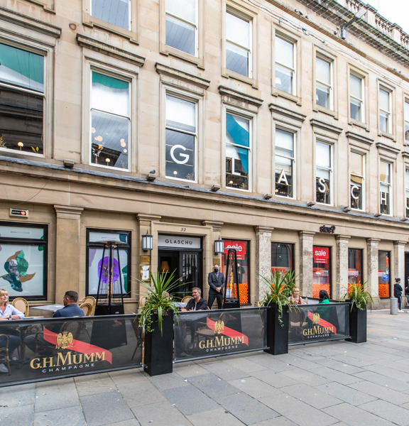 An exterior image of Glaschu's entrance. A grand, blonde sandstone building over 3 storey's, beside a paved pedestrianised area. Planters and banners separate a small private area of outdoor seating from the walkway to the left of the entrance, diners sit at 2 of the tables, either side of a stylish space heater. The first story windows of the building are decorated with colourful vinyls in organic "blobby" shapes. A white and gold letter sits prominently in each window, reading "GLASCHU".