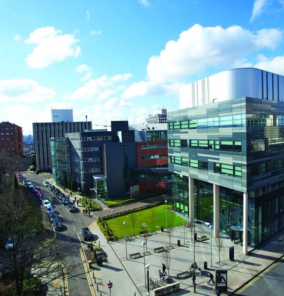 A photo overlooking buildings on the University of Strathclyde campus from a high-up vantage point. A glass and silver, modern, square building sits on a junction between 2 roads. A large paved and landscaped area with benches and bins sits directly in front of it, adjoining the wide pavement that wraps around the street corner. Other modern buildings with colourful cladding and older brick buildings can be seen on either side of the surrounding roads. Parked cars and pedestrians can also be seen.