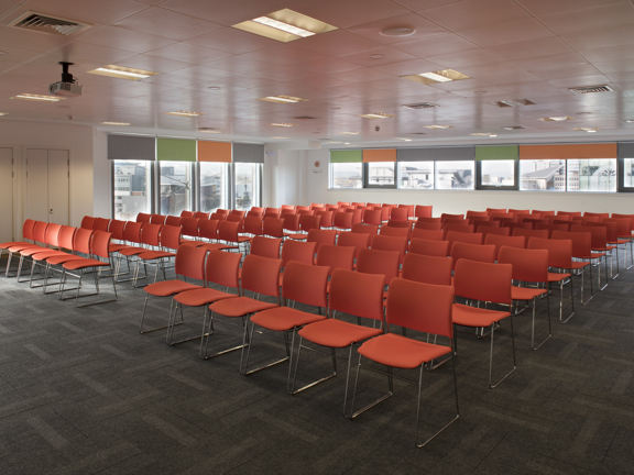 A meeting set up at The Studio shows the corner of a large room with white walls and dark grey carpet tiles. Windows with different coloured roller blinds, in orange, grey and green, wrap around the corner of the room. Dozens of red fabric chairs with wire legs are arranged into 9 rows with a central aisle. A projector is mounted on the tiled ceiling, between fluorescent lights. 