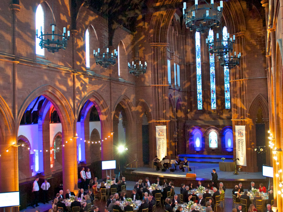 Strathclyde University's ceremonial Barony Hall decorated & in use for a university dinner. The building, a gothic, red-sandstone, former church is cavernous - with towering carved stone arches and a vaulted wooden ceiling. It is possible to see the floor is stone but a large carpet has been placed in the main space. Banners and fairy lights decorate the naves pillars. Dozens of round tables, decorated with candles and flowers are each occupied by 10 guests. 4 large TV screens on stands are also visible.