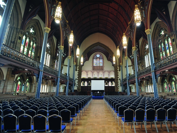 The UoG's Bute Hall arranged for a lecture or talk.  A magnificent space with wooden floors, stained glass windows & a tall 2-storey vaulted, wooden ceiling. A first floor balcony can be seen running the length of the outer walls. Blue velvet chairs with metal legs are arranged into dozens of rows with a wide central isle. Carved, wooden railings & benches can be seen around the rooms edges, on both floors. A temporary stage with blue carpeting, a panel of chairs and a projection screen can also be seen.