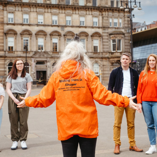 Woman wearing orange jacket with Walking Tours of Glasgow on the back speaking to 4 people