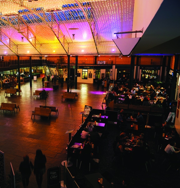 An interior view of Merchant Square, shows a large open space with a paved stone floor and a curving roof. The photo is taken from a first floor balcony at night, a mesh of fairy lights covers the ceiling and spotlights also light the tops of the walls. The open space has a dozen wooden benches placed back to back. Around the open space, fenced off seating areas and the entrances to several businesses are visible. A glass lift between the ground floor and the first floor balcony can be seen on the left.