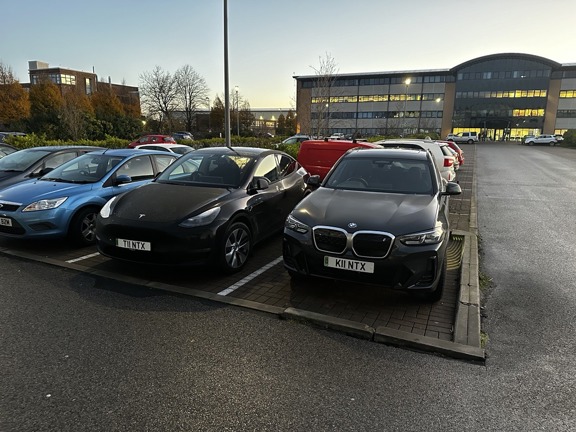Several cars parked in a large carpark, in the foreground an electric BMW and a Tesla are visible, both cars are black. The picture is taken in the evening and modern office buildings can be seen lit at the far end of the car park are beyond some trees to the left.
